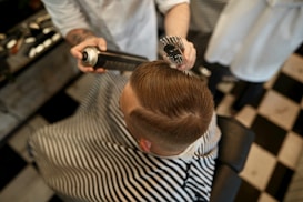 A person is sitting in a barber chair, covered with a striped cape. The barber is styling the person's hair using a round brush and a can of hair spray. The setting appears to be a barbershop with a black and white checkered floor.