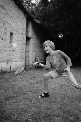 A black and white image of a young boy playing badminton outdoors. The boy is focused and mid-action as he strikes the shuttlecock with a racket. He is wearing a long-sleeved shirt, shorts, and sandals. The setting appears rustic, with a stone building and trees in the background.