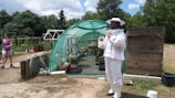 A person in a beekeeping suit is standing outside a green mesh greenhouse, with various potted plants and gardening tools inside. To the left, another person in casual clothing walks on a dirt path. The scene is set in a lush, green environment with trees and cloudy skies in the background.