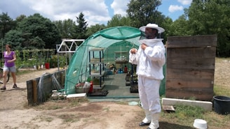 A person in a beekeeping suit is standing outside a green mesh greenhouse, with various potted plants and gardening tools inside. To the left, another person in casual clothing walks on a dirt path. The scene is set in a lush, green environment with trees and cloudy skies in the background.