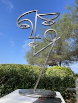 A metal sculpture represents a stylized human face with abstract lines and geometric features. It stands outdoors with a backdrop of green shrubs and a tree, all under a clear blue sky.