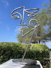 A metal sculpture represents a stylized human face with abstract lines and geometric features. It stands outdoors with a backdrop of green shrubs and a tree, all under a clear blue sky.