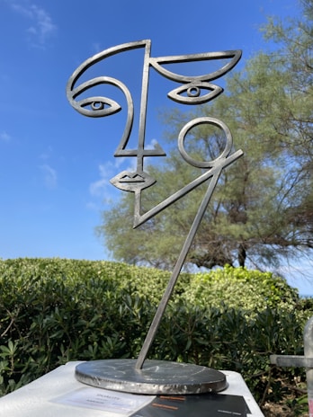 A metal sculpture represents a stylized human face with abstract lines and geometric features. It stands outdoors with a backdrop of green shrubs and a tree, all under a clear blue sky.