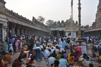 A large group of people gathered in an ancient temple courtyard, engaging in a religious ceremony. Many individuals are lighting small clay lamps and placing them on the ground. The architecture is richly detailed with numerous columns and carvings, and the scene is lively with movement and color.