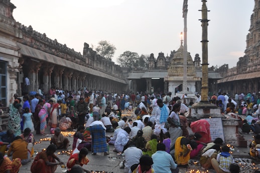 A large group of people gathered in an ancient temple courtyard, engaging in a religious ceremony. Many individuals are lighting small clay lamps and placing them on the ground. The architecture is richly detailed with numerous columns and carvings, and the scene is lively with movement and color.