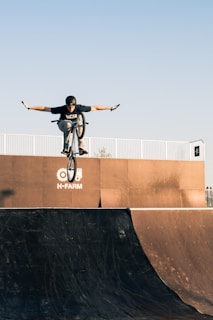 A person is performing a stunt on a BMX bike, flying through the air with arms outstretched. The scene takes place on a skateboard ramp, with a clear blue sky in the background. The ramp has a logo that reads 'H-FARM'. The rider is wearing a helmet and casual clothing.