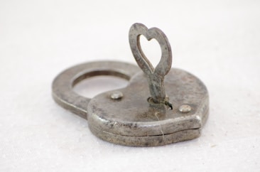 An antique metal lock shaped like a heart with a keyhole in the center. The lock is old and slightly tarnished, featuring a heart-shaped key inserted into its top.
