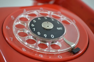 Close-up of a vintage red rotary dial telephone with clear plastic finger wheel and black center displaying numbers. The metal finger stop and a portion of the phone's body are visible.