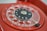 Close-up of a vintage red rotary dial telephone with clear plastic finger wheel and black center displaying numbers. The metal finger stop and a portion of the phone's body are visible.