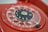 Close-up of a vintage red rotary dial telephone with clear plastic finger wheel and black center displaying numbers. The metal finger stop and a portion of the phone's body are visible.