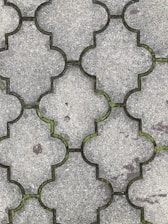 Interlocking concrete tiles create a geometric pattern on a walkway. The tiles have a light gray color, and small patches of green moss grow in the gaps between them. The surface of the tiles appears worn, with slight discolorations and spots visible.