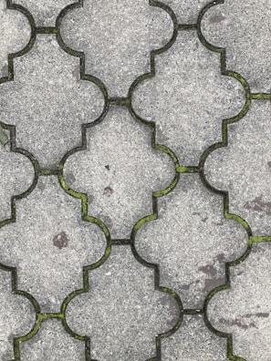 Interlocking concrete tiles create a geometric pattern on a walkway. The tiles have a light gray color, and small patches of green moss grow in the gaps between them. The surface of the tiles appears worn, with slight discolorations and spots visible.