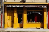 A charming storefront with a bright yellow facade featuring wooden doors and window frames. The awning at the top is dark brown with white lettering that reads 'Cordonnerie Traditionnelle - Clés.' Inside the window display, there are various shoes and cobbler tools visible against a background of red curtains. The street in front has a grey sidewalk with a yellow line at the curb.