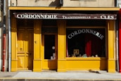 A charming storefront with a bright yellow facade featuring wooden doors and window frames. The awning at the top is dark brown with white lettering that reads 'Cordonnerie Traditionnelle - Clés.' Inside the window display, there are various shoes and cobbler tools visible against a background of red curtains. The street in front has a grey sidewalk with a yellow line at the curb.