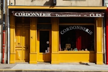 A charming storefront with a bright yellow facade featuring wooden doors and window frames. The awning at the top is dark brown with white lettering that reads 'Cordonnerie Traditionnelle - Cl&eacute;s.' Inside the window display, there are various shoes and cobbler tools visible against a background of red curtains. The street in front has a grey sidewalk with a yellow line at the curb.
