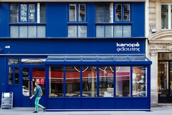 A vibrant blue storefront with large windows and a sign displaying 'kanop&eacute;' above. The building has a modern and clean design with contrasting elements such as wood and glass. A person in colorful clothing is walking past, adding a dynamic element to the scene. Several chairs and a table can be seen through the windows, hinting at a caf&eacute; or restaurant setting.
