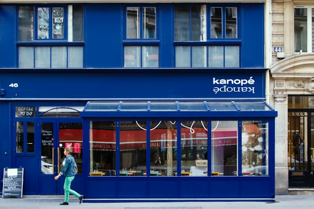 A vibrant blue storefront with large windows and a sign displaying 'kanop&eacute;' above. The building has a modern and clean design with contrasting elements such as wood and glass. A person in colorful clothing is walking past, adding a dynamic element to the scene. Several chairs and a table can be seen through the windows, hinting at a caf&eacute; or restaurant setting.