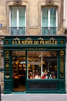 A charming, old-fashioned storefront with the name 'À LA MÈRE DE FAMILLE' displayed prominently in gold letters on a dark green facade. The store features large glass windows showcasing various goods inside. The building's architecture consists of ornate details and two large windows on the floor above the shop. There are decorative wrought iron designs under the windows.