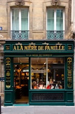 A charming, old-fashioned storefront with the name 'À LA MÈRE DE FAMILLE' displayed prominently in gold letters on a dark green facade. The store features large glass windows showcasing various goods inside. The building's architecture consists of ornate details and two large windows on the floor above the shop. There are decorative wrought iron designs under the windows.