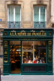 A charming, old-fashioned storefront with the name 'À LA MÈRE DE FAMILLE' displayed prominently in gold letters on a dark green facade. The store features large glass windows showcasing various goods inside. The building's architecture consists of ornate details and two large windows on the floor above the shop. There are decorative wrought iron designs under the windows.