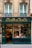 A charming, old-fashioned storefront with the name 'À LA MÈRE DE FAMILLE' displayed prominently in gold letters on a dark green facade. The store features large glass windows showcasing various goods inside. The building's architecture consists of ornate details and two large windows on the floor above the shop. There are decorative wrought iron designs under the windows.