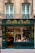 A charming, old-fashioned storefront with the name 'À LA MÈRE DE FAMILLE' displayed prominently in gold letters on a dark green facade. The store features large glass windows showcasing various goods inside. The building's architecture consists of ornate details and two large windows on the floor above the shop. There are decorative wrought iron designs under the windows.