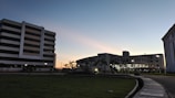 A modern campus is depicted at dusk with several multi-story buildings. The sky displays a gradient of colors from blue to orange along the horizon. Lights illuminate pathways and the interiors of buildings, while palm trees line the roundabout. The foreground features a neatly maintained lawn, and a curved pavement extends into the composition.