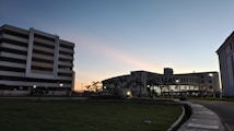 A modern campus is depicted at dusk with several multi-story buildings. The sky displays a gradient of colors from blue to orange along the horizon. Lights illuminate pathways and the interiors of buildings, while palm trees line the roundabout. The foreground features a neatly maintained lawn, and a curved pavement extends into the composition.