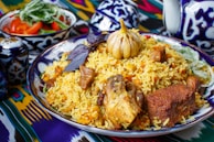 A plate of vibrant and colorful rice dish with chunks of meat, a whole bulb of garlic, and garnishments such as herbs on top. The plate is intricately decorated with patterns and surrounded by similarly styled tableware. A small bowl with fresh tomatoes and greens is visible in the background.