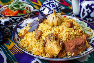 A plate of vibrant and colorful rice dish with chunks of meat, a whole bulb of garlic, and garnishments such as herbs on top. The plate is intricately decorated with patterns and surrounded by similarly styled tableware. A small bowl with fresh tomatoes and greens is visible in the background.