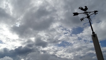 A weather vane featuring a rooster silhouette stands against a backdrop of a cloudy sky. The clouds are thick and varied in shades of gray, with subtle hints of blue peeking through.