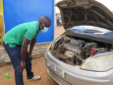 A person wearing a green shirt and a face mask is inspecting the engine of a car with the hood open. The car is parked on a dirt ground near a blue wall and a yellow sign with text. A green bottle is lying on the ground next to the person.