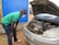 A person wearing a green shirt and a face mask is inspecting the engine of a car with the hood open. The car is parked on a dirt ground near a blue wall and a yellow sign with text. A green bottle is lying on the ground next to the person.