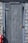 A weathered, rustic wooden door set into a shingled wall. The door is painted with a faded blue-gray color, and there is a red and white buoy hanging on the left side. The metal hinge and latch on the door appear aged.