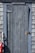 A weathered, rustic wooden door set into a shingled wall. The door is painted with a faded blue-gray color, and there is a red and white buoy hanging on the left side. The metal hinge and latch on the door appear aged.