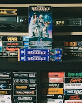 A vintage collection of VHS tapes displayed on shelves, featuring titles such as 'Beetlejuice', 'Halloween', and 'Night of the Living Dead'. The focus is on the 'Beetlejuice' tapes prominently stacked in the center. A VHS player and a digital clock displaying 3:29 are seen in the background, surrounded by a variety of horror and thriller movie tapes, contributing to a nostalgic atmosphere.