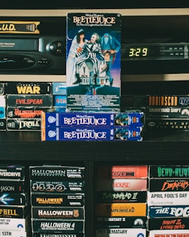 A vintage collection of VHS tapes displayed on shelves, featuring titles such as 'Beetlejuice', 'Halloween', and 'Night of the Living Dead'. The focus is on the 'Beetlejuice' tapes prominently stacked in the center. A VHS player and a digital clock displaying 3:29 are seen in the background, surrounded by a variety of horror and thriller movie tapes, contributing to a nostalgic atmosphere.