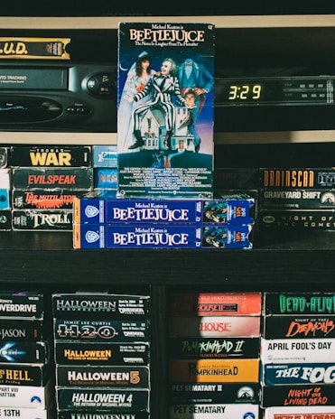 A vintage collection of VHS tapes displayed on shelves, featuring titles such as 'Beetlejuice', 'Halloween', and 'Night of the Living Dead'. The focus is on the 'Beetlejuice' tapes prominently stacked in the center. A VHS player and a digital clock displaying 3:29 are seen in the background, surrounded by a variety of horror and thriller movie tapes, contributing to a nostalgic atmosphere.