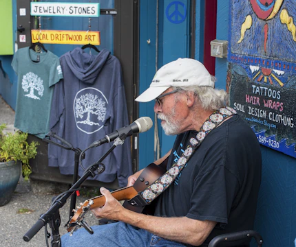 An older man with a white beard and white cap is playing an acoustic guitar and singing into a microphone. He is seated on a chair outside a shop displaying signs and merchandise. The shop has colorful signs reading 'Jewelry-Stones' and 'Local Driftwood Art,' and displays of clothing with tree designs. A painted sign advertises tattoos and soul design clothing. There is a blue wall, suggesting a laid-back, artistic atmosphere.