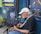 An older man with a white beard and white cap is playing an acoustic guitar and singing into a microphone. He is seated on a chair outside a shop displaying signs and merchandise. The shop has colorful signs reading 'Jewelry-Stones' and 'Local Driftwood Art,' and displays of clothing with tree designs. A painted sign advertises tattoos and soul design clothing. There is a blue wall, suggesting a laid-back, artistic atmosphere.