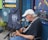 An older man with a white beard and white cap is playing an acoustic guitar and singing into a microphone. He is seated on a chair outside a shop displaying signs and merchandise. The shop has colorful signs reading 'Jewelry-Stones' and 'Local Driftwood Art,' and displays of clothing with tree designs. A painted sign advertises tattoos and soul design clothing. There is a blue wall, suggesting a laid-back, artistic atmosphere.