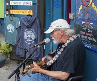 An older man with a white beard and white cap is playing an acoustic guitar and singing into a microphone. He is seated on a chair outside a shop displaying signs and merchandise. The shop has colorful signs reading 'Jewelry-Stones' and 'Local Driftwood Art,' and displays of clothing with tree designs. A painted sign advertises tattoos and soul design clothing. There is a blue wall, suggesting a laid-back, artistic atmosphere.