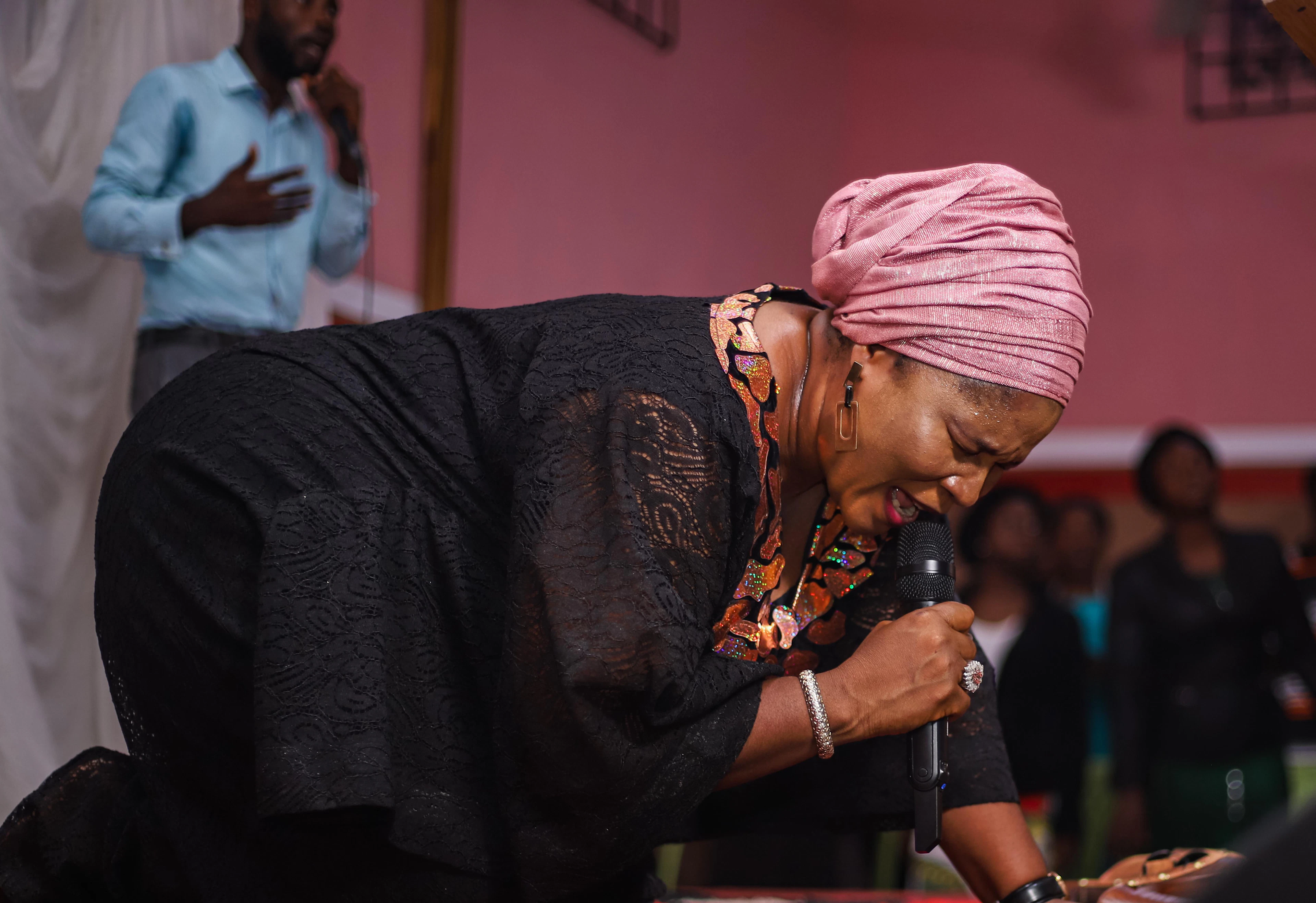 An older woman in a pink head wrap speaking into a handheld microphone