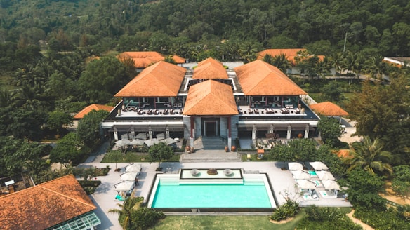 A large resort-style building with several terracotta roofs surrounded by lush green trees and foliage. In front of the building, there is a rectangular swimming pool with sun loungers and umbrellas on a stone patio. The setting is tropical, with palm trees visible around the property, and a forested hillside in the background.