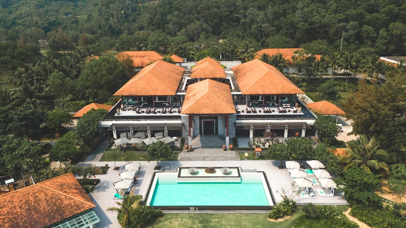A large resort-style building with several terracotta roofs surrounded by lush green trees and foliage. In front of the building, there is a rectangular swimming pool with sun loungers and umbrellas on a stone patio. The setting is tropical, with palm trees visible around the property, and a forested hillside in the background.
