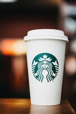 A white disposable coffee cup with a plastic lid is prominently displayed, featuring a green logo with a twin-tailed mermaid at the center. The cup is resting on a wooden surface with a blurred, warm-toned background.