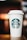A white disposable coffee cup with a plastic lid is prominently displayed, featuring a green logo with a twin-tailed mermaid at the center. The cup is resting on a wooden surface with a blurred, warm-toned background.