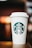 A white disposable coffee cup with a plastic lid is prominently displayed, featuring a green logo with a twin-tailed mermaid at the center. The cup is resting on a wooden surface with a blurred, warm-toned background.