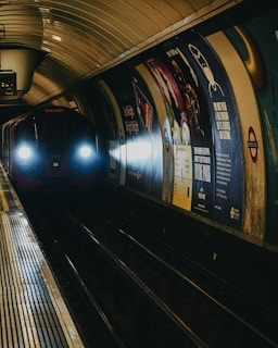 A subway train approaches a dimly lit underground station. The tunnel features advertisements on the walls, and the train's headlights illuminate the tracks. The environment appears slightly moist with a metallic sheen on the platform.
