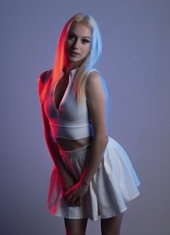 A woman with light blonde hair stands posing in a studio setting, illuminated by vibrant red and blue lighting. She is wearing a white sleeveless crop top and a white skirt. The contrasting lighting creates a dramatic effect on her hair and outfit.